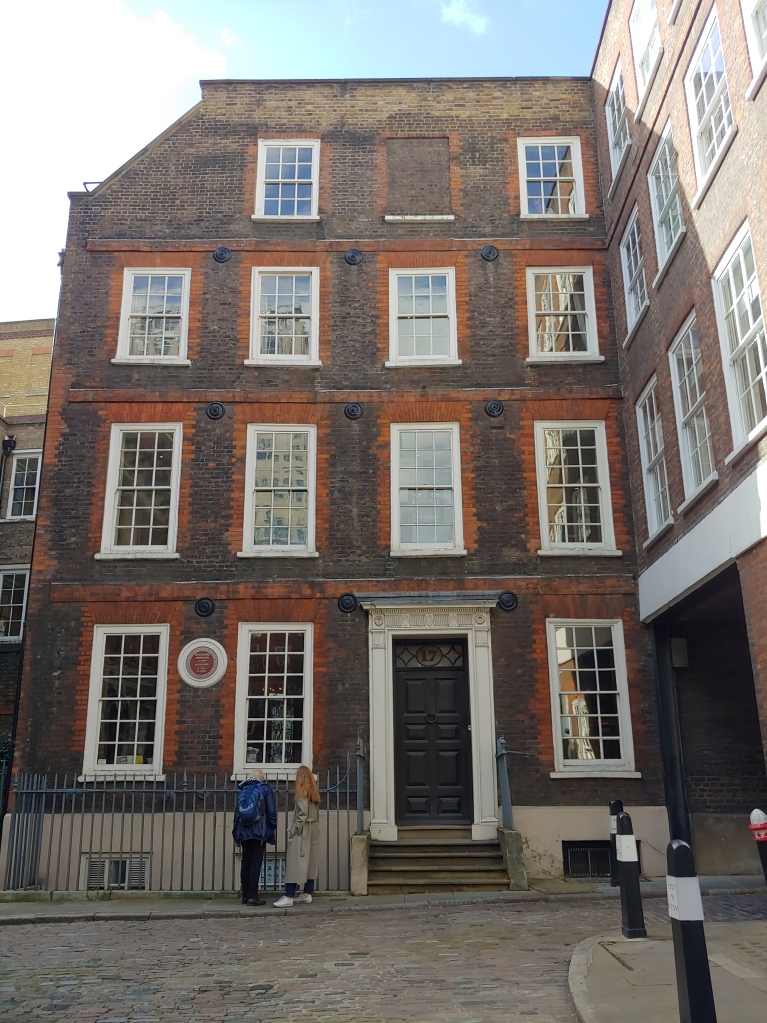 Image of red brick house with white sash windows tucked away in the corner of a square, with a large black door.