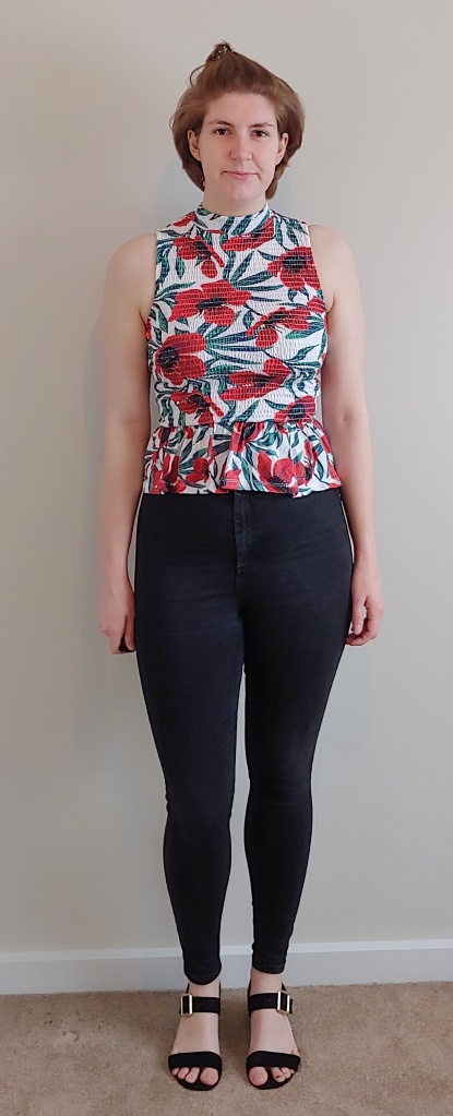 Full length photo of Helen wearing a poppy print peplum top with mock neck, over black jeans with black sandals.