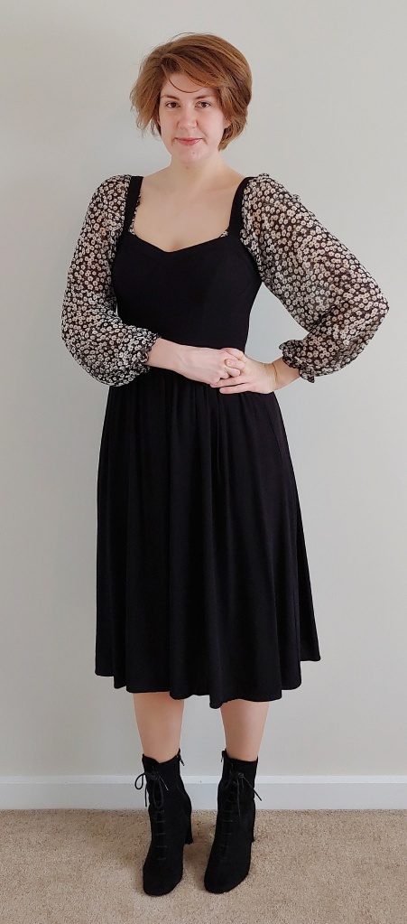 Full length photo of Helen wearing a black pinafore dress with a black, white, and yellow floral floaty top underneath.