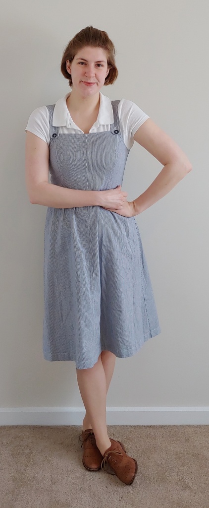 Full length photo of Helen wearing a knee-length pinafore dress in thin white and blue vertical stripes, with a white short-sleeved polo shirt underneath, and brown brogues.