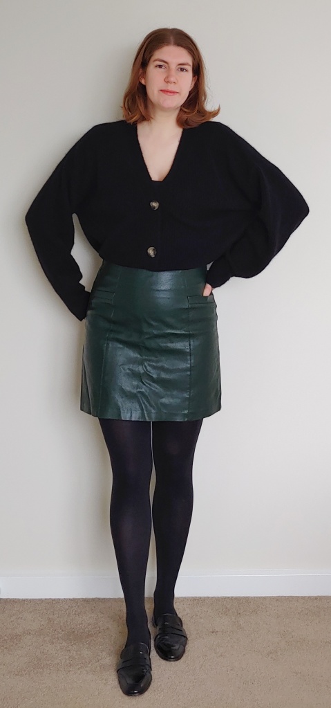 Helen wearing a short green leather-look skirt with black tights and loafers, with a thick black cardigan.