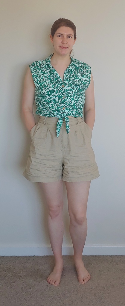 Full length photo of Helen wearing A-line beige high-waisted shorts with a sleeveless, button-through and tie-waist shirt in a turquoise-green on white leaf print.