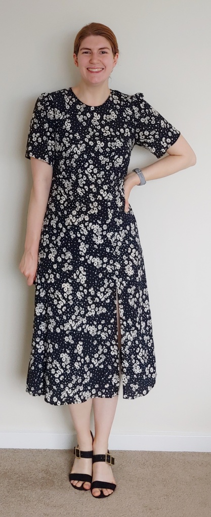 Full-length photo of Helen wearing a round-neck midi dress with a side slit and short sleeves. It is black with daisy-like flowers with white petals and yellow centres printed on it. She also wears black, block-heeled sandals.
