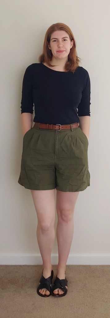Helen wearing half-sleeved black boat-neck t-shirt and high-waisted khaki shorts, with a brown belt and black cross-over sandals.