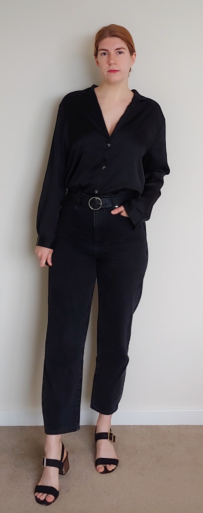 Full length photo of Helen wearing wide-leg black jeans with a black long-sleeved shirt with tortoiseshell buttons, and black belt with circular buckle, and black block-heeled sandals.