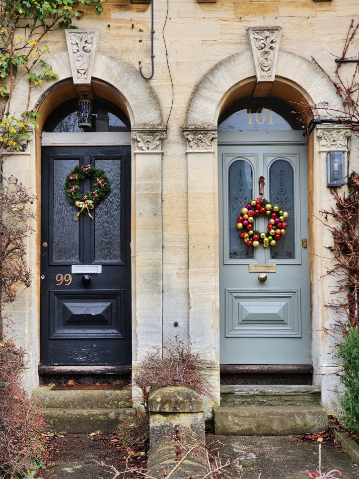 Another pair of Victorian doors, this time with rounded archways above, which each feature a decorative carving with their house numbers in, which are different to the modern numbers on the doors. The door on the left is black with a traditional wreath of green with some red highlights and a gold coloured bow at the bottom, while the door on the right is a sage-grey colour with a wreath of brightly coloured gold, orange, pink and green baubles on a base of fir.