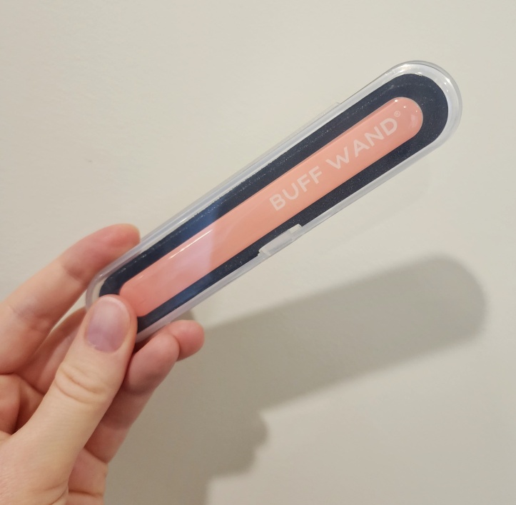 Left hand holding a small plastic case, with a pink emery-board shaped file with 'BUFF WAND' written on it in all capitals.